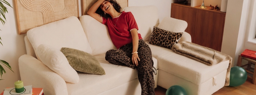 Femme dans un salon cosy avec canapé blanc et coussins imprimés Femme assise sur un canapé d'angle blanc avec coussins décoratifs, plaid et mobilier en bois, ballons au sol et décoration murale en relief