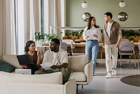 Deux personnes assises sur un canapé discutant avec documents et ordinateur portable, tandis que deux autres marchent dans un bureau moderne avec grandes baies vitrées et mobilier design