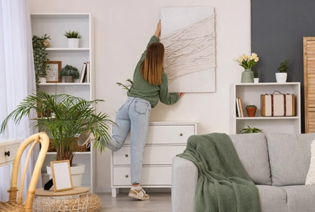 Personne accrochant un tableau abstrait au-dessus d'une commode blanche dans un salon décoré avec plantes vertes, canapé gris et étagères